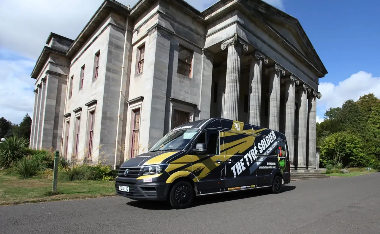 The Tyre Soldier black VW Crafter van, gold tribal-stripe livery, parked in front of a grand columned Georgian estate building at Camperdown, Dundee.